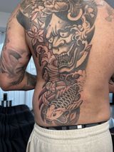 Person showing a large black-and-gray back tattoo of a fierce mask and koi fish, seen from behind.