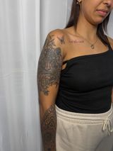 Woman in a black one-shoulder top and beige pants, showing tattooed arm against a light curtain.