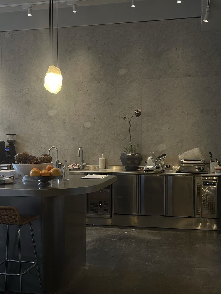 Modern kitchen with stainless steel island and countertops, gray concrete walls, and hanging light fixture.
