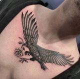 Black bird tattoo with wings spread, carrying a rose on a person’s shoulder near the neck.