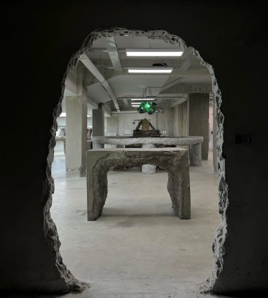 A view through a damaged doorway into a gallery. Gray concrete tables are displayed. Bright overhead lights.
