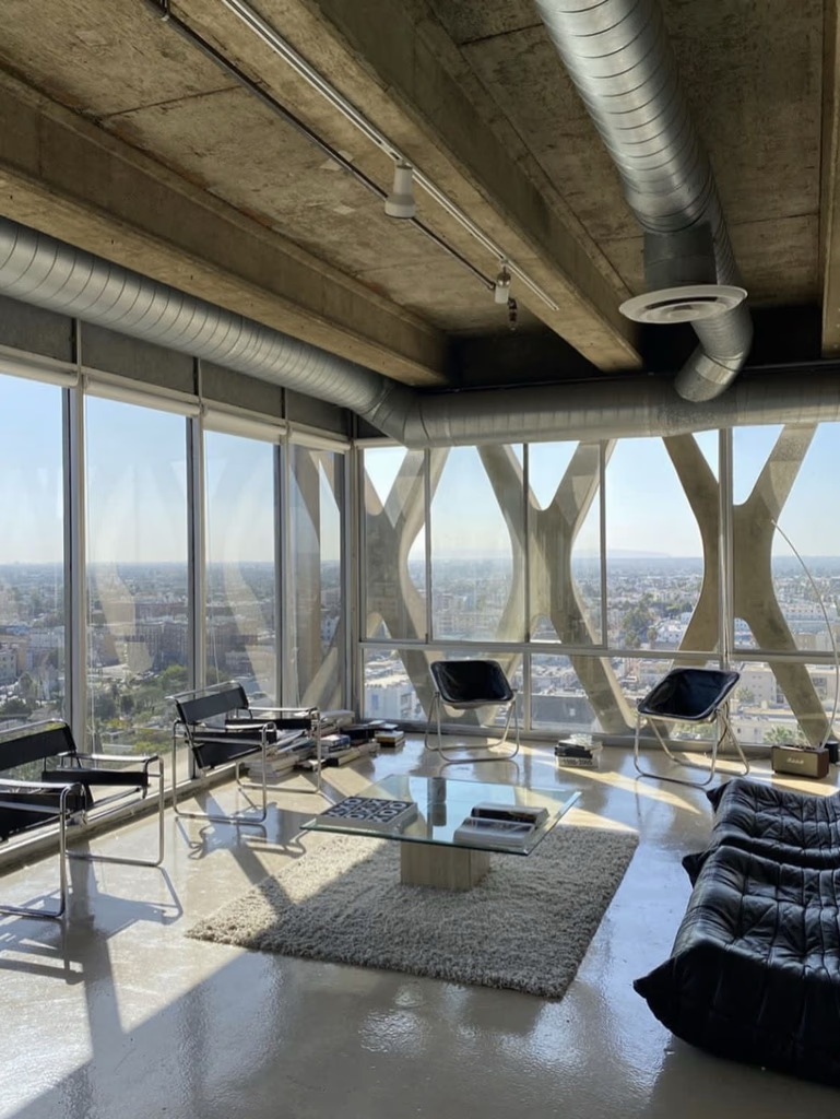 Modern living room with city view, large windows, sleek furniture, and concrete ceiling.