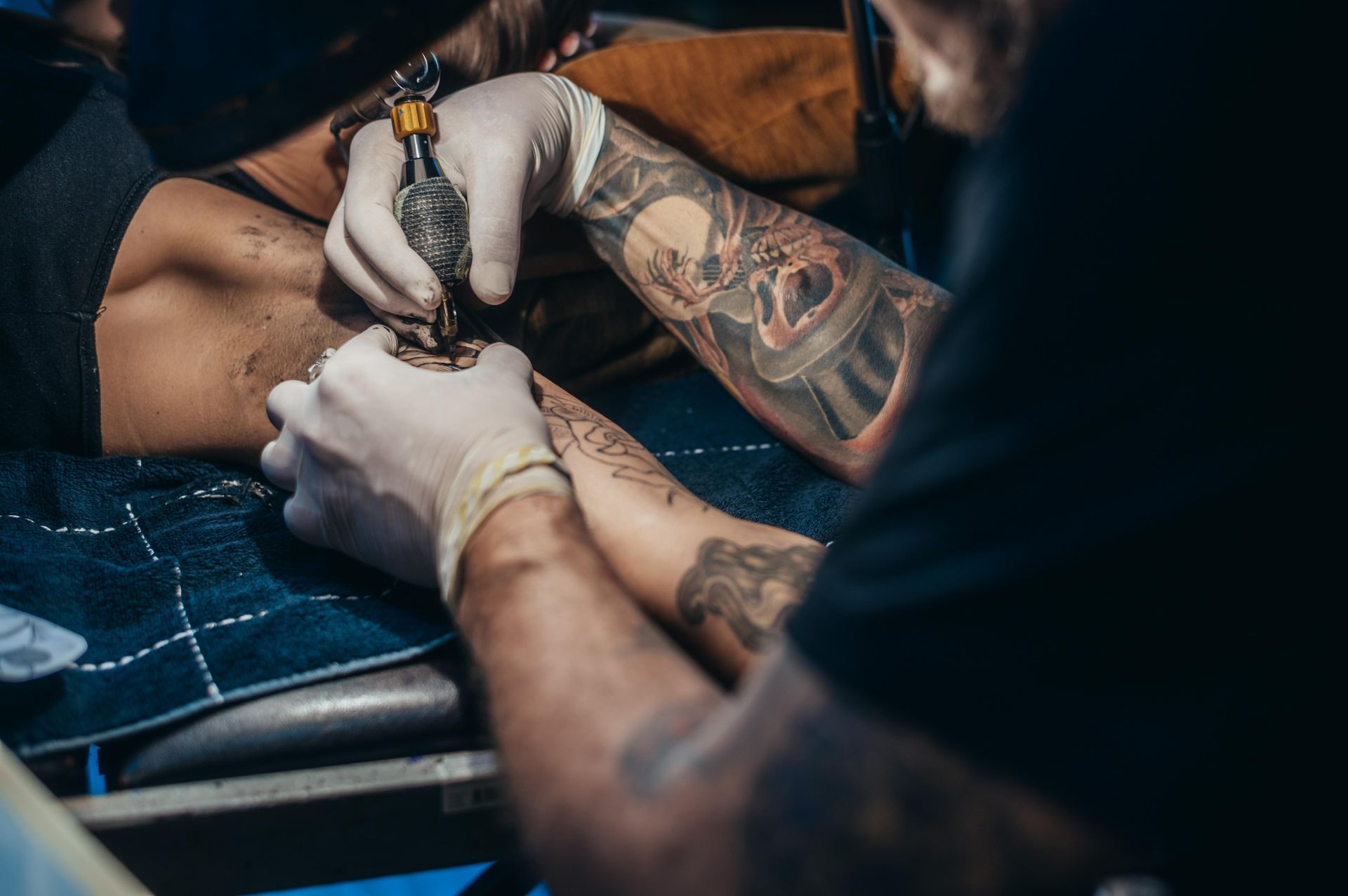 Person Getting A Tattoo On Their Arm From An Artist Wearing Gloves; Blue Table, Dark Lighting — Tattoo Inc Studio in Burleigh, QLD