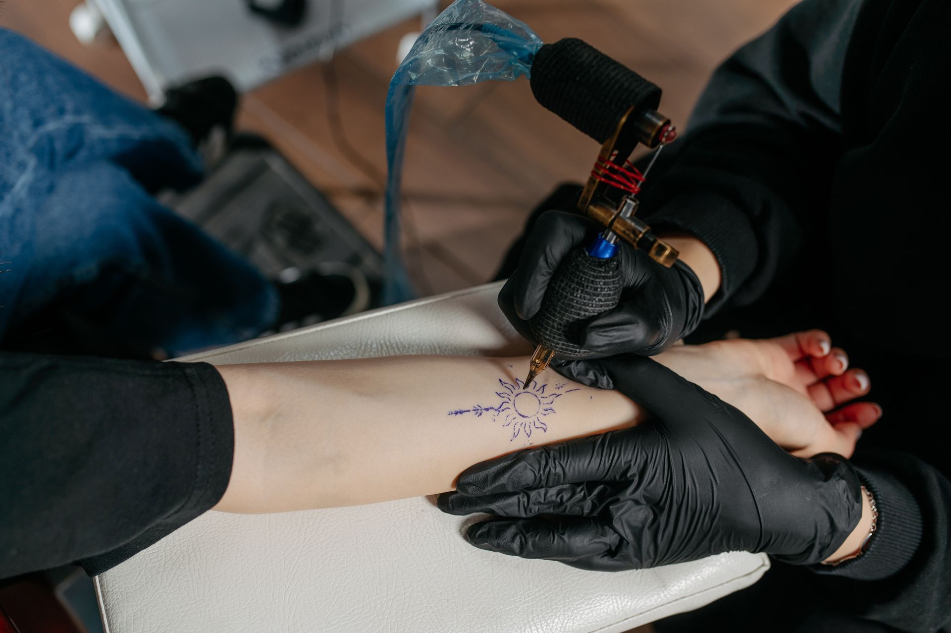 Tattoo Artist In Black Gloves Tattooing A Blue Design On A Person's Wrist — Tattoo Inc Studio in Robina, QLD