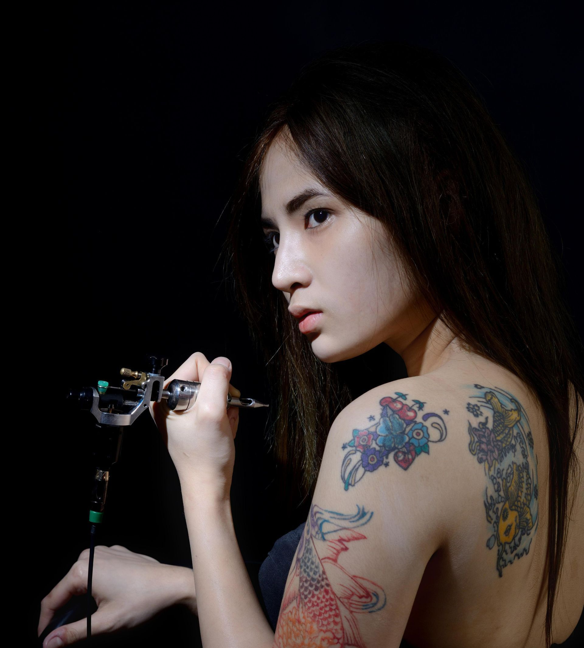 Woman With Tattoos Holds Tattoo Gun, Looking To The Side Against A Black Background — Tattoo Inc Studio in Burleigh, QLD
