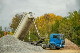Blue dump truck unloading gravel on a pile, outdoors with fall foliage in background.