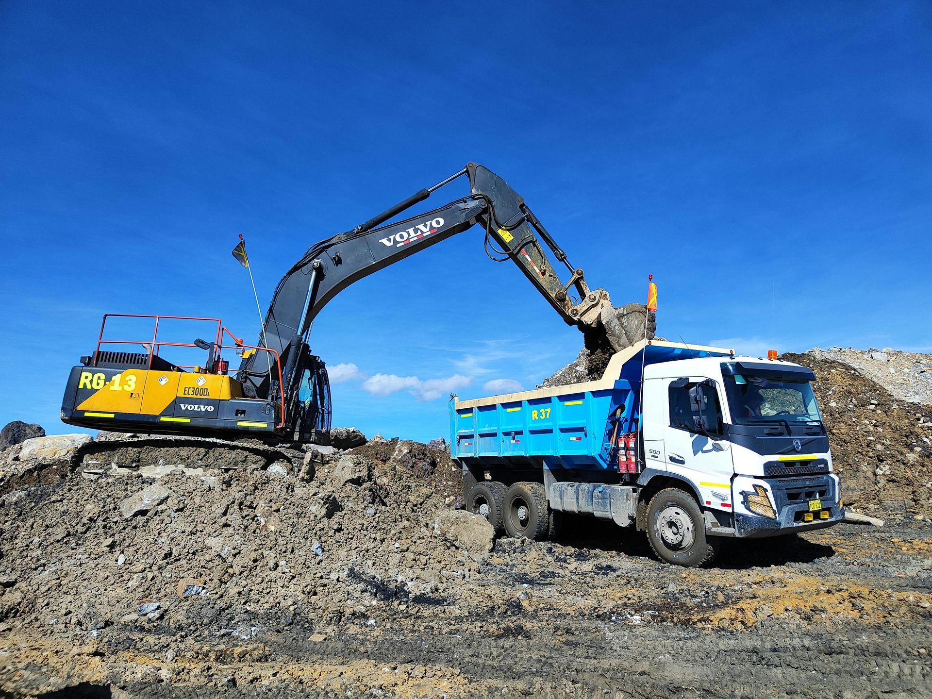 Yellow and black excavator loading a blue-dump truck with dirt under a blue sky.