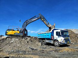 Yellow and black excavator loading a blue-dump truck with dirt under a blue sky.