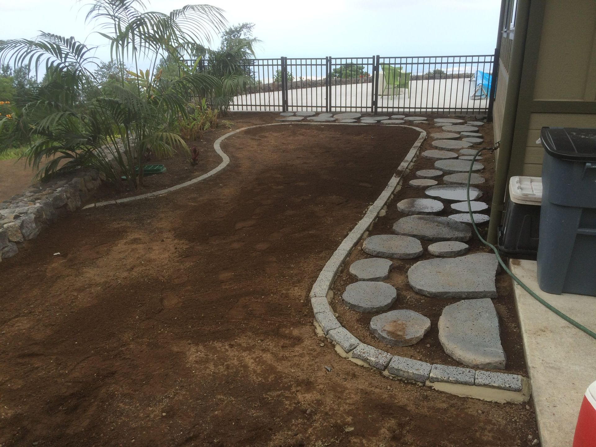 A stone pathway curves through a mulched garden, next to a building, with a gated fence in the background.
