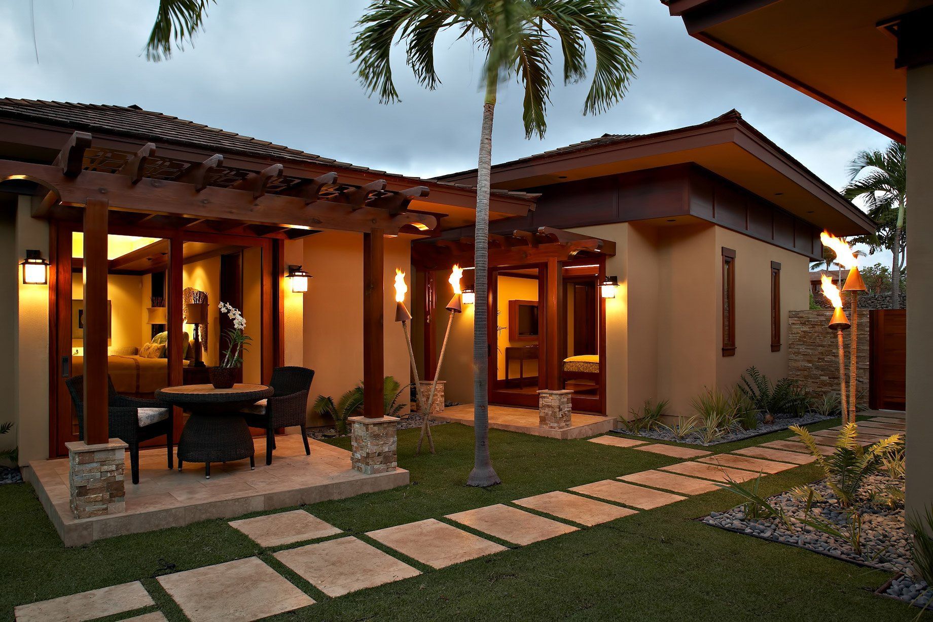 Tropical house exterior with patio, pathway, palm tree, and tiki torches at dusk.