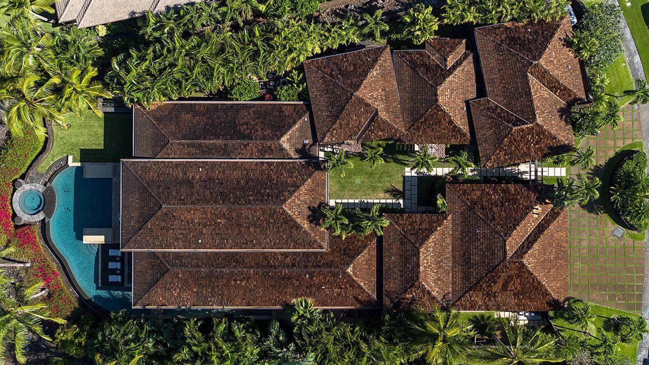 Aerial view of a large brown-roofed house with a pool and lush greenery surrounding it.