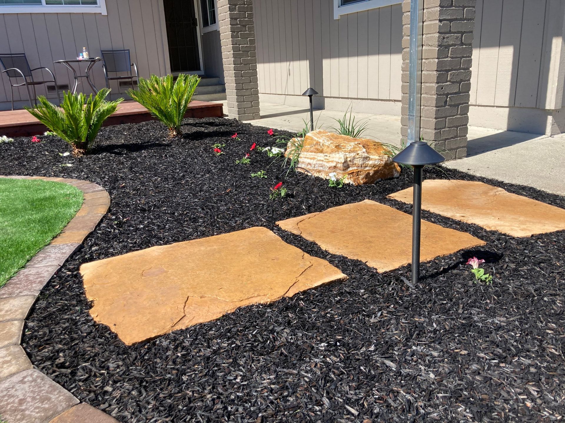 Black mulched flowerbed with stepping stones, plants, a rock, and a pathway.