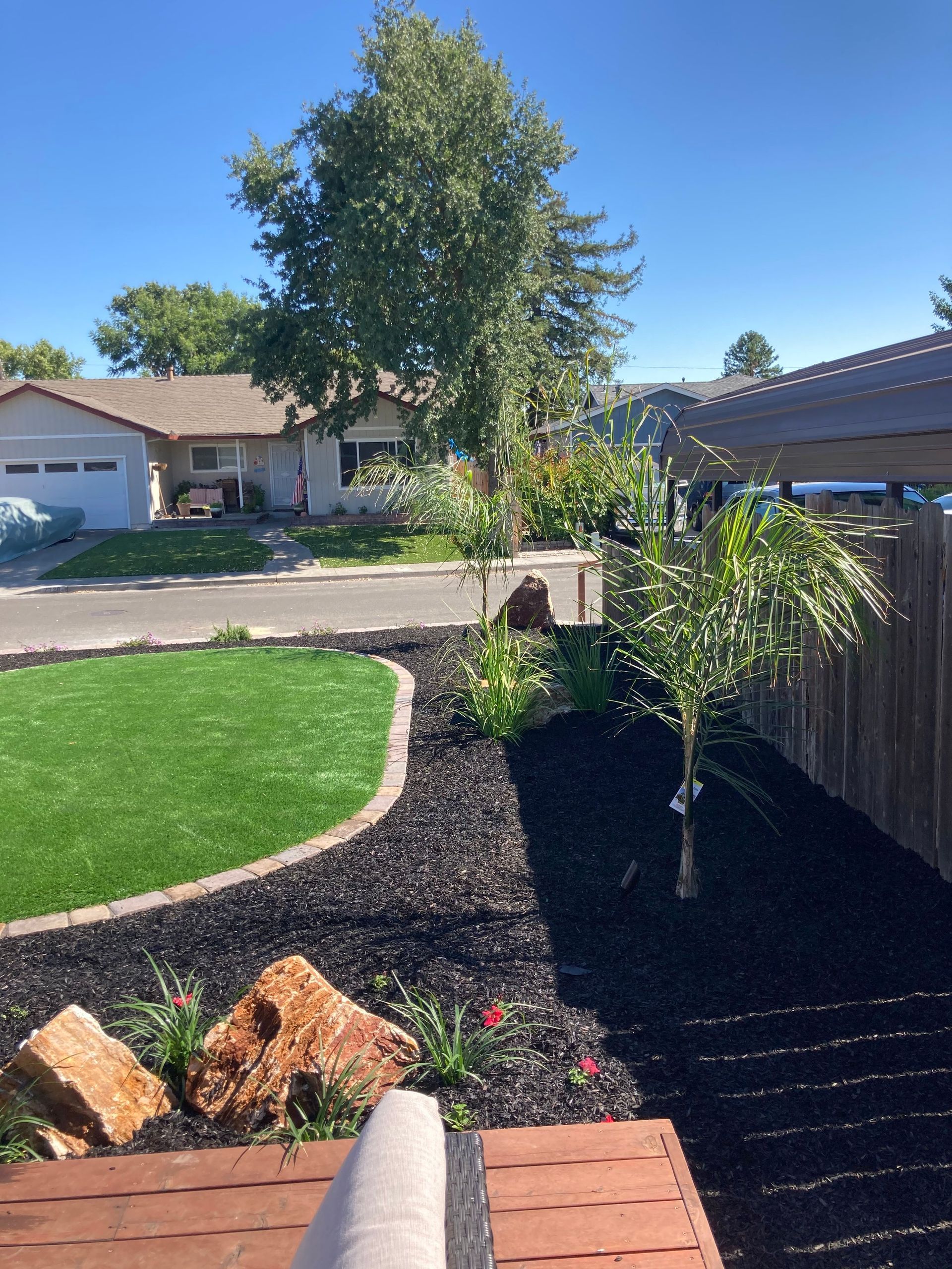 Backyard with artificial turf, black mulch, small tree, rocks, and wooden deck. Blue sky, sunny day.