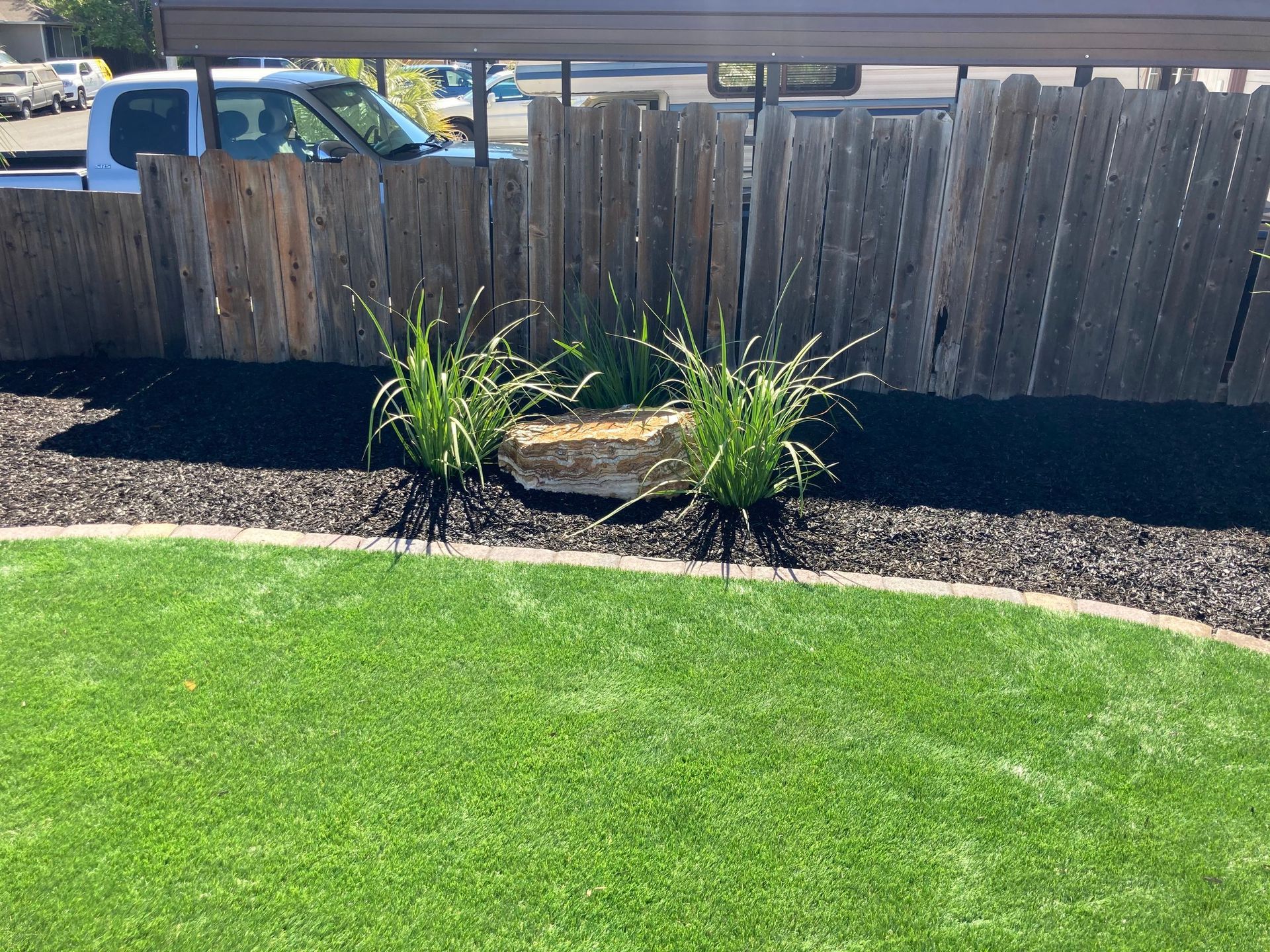 Lush green grass with a small garden bed of black mulch, plants, and a large rock in front of a wooden fence.