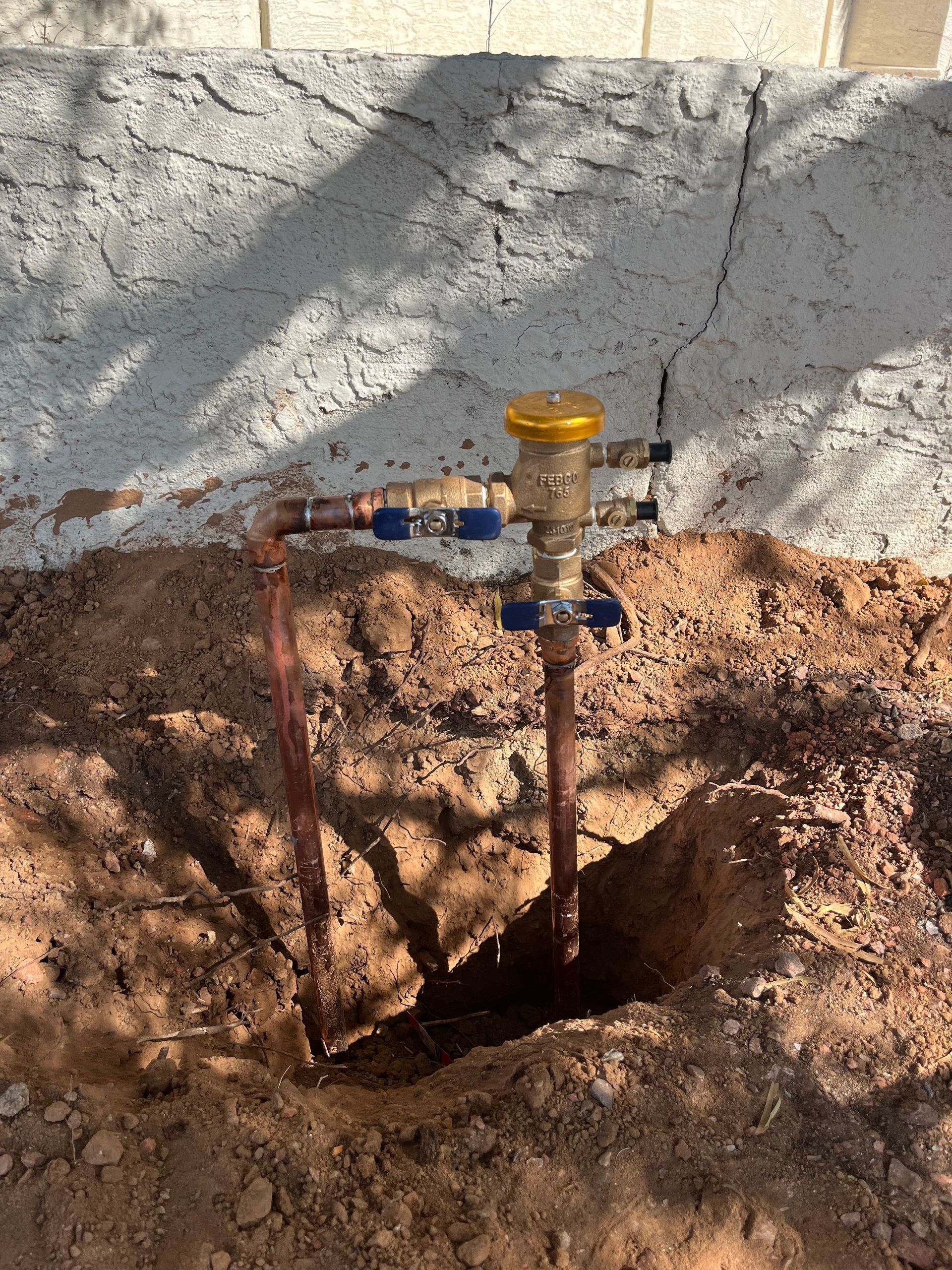 Copper pipes and sprinkler valve emerging from a hole in the dirt near a textured wall.