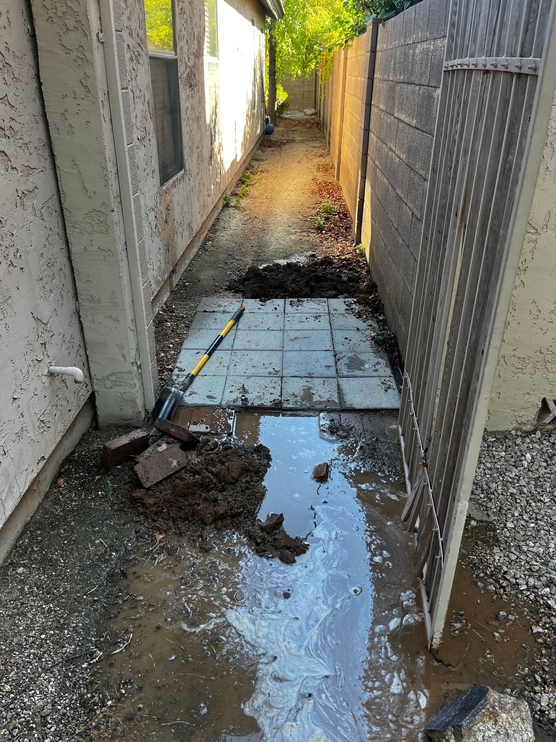 Muddy pathway between two walls; water puddle, dirt, tiles, and a gardening tool present.