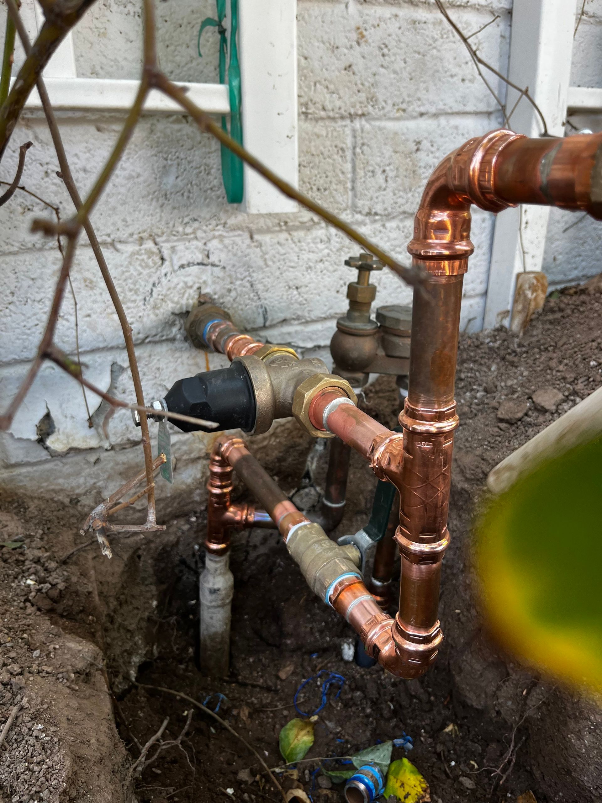 Copper pipes and fittings are exposed in a dug-out trench next to a light brick wall.