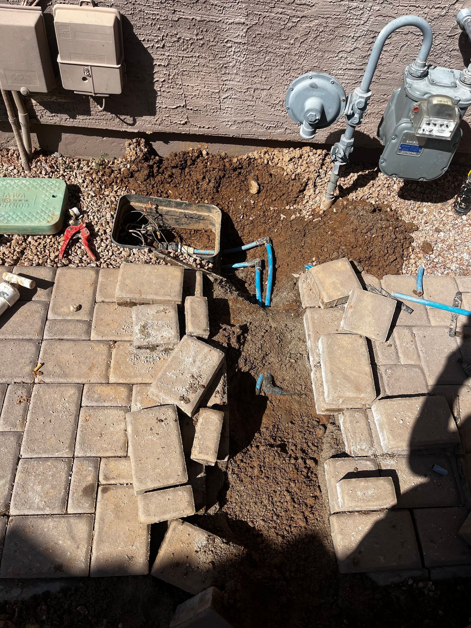 Brick patio partially dug up near a house. Exposed dirt, a blue pipe, and a gas meter are visible.
