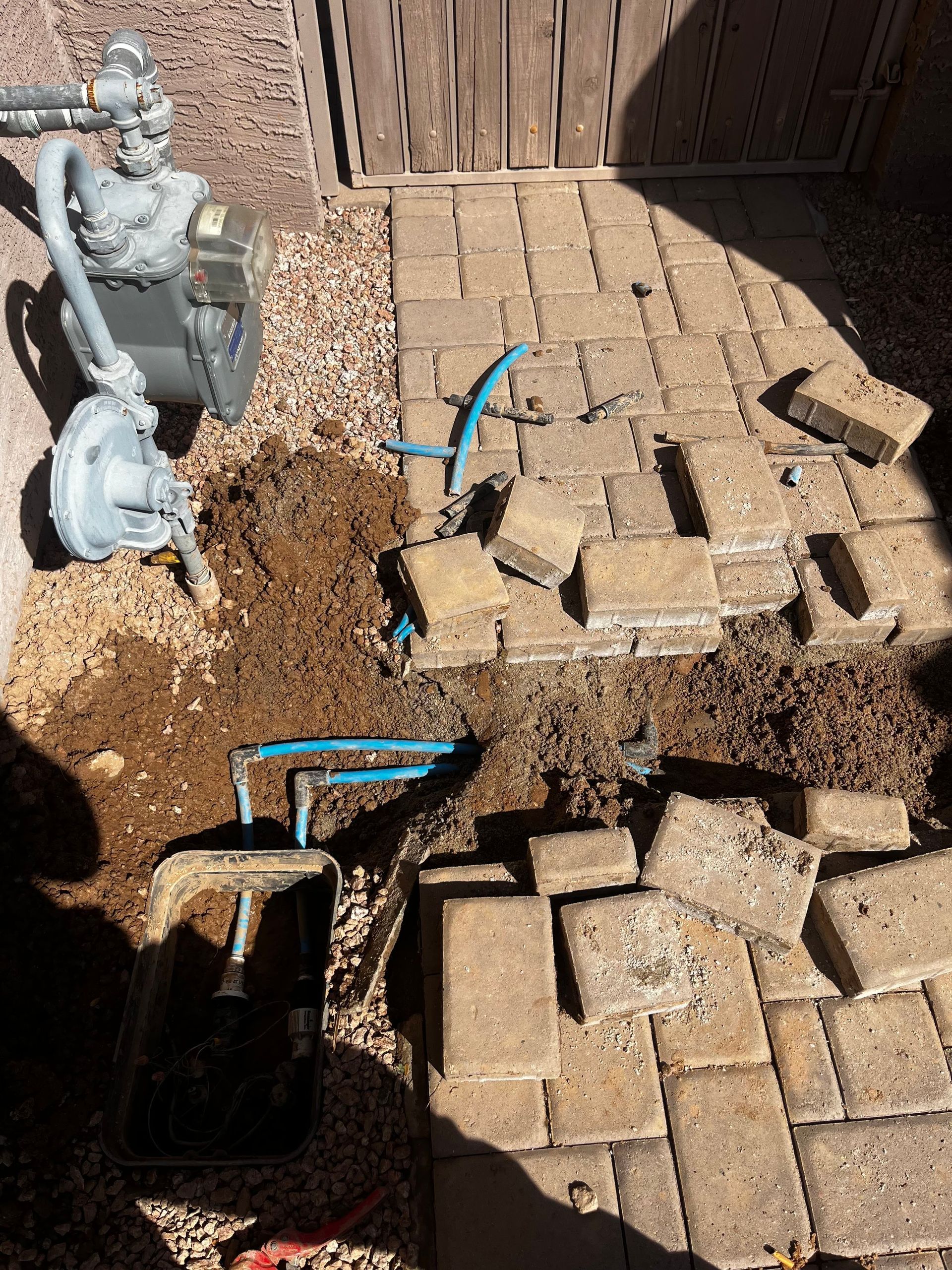 Patio pavers removed for plumbing work near a gas meter. Brown dirt and blue pipes visible.