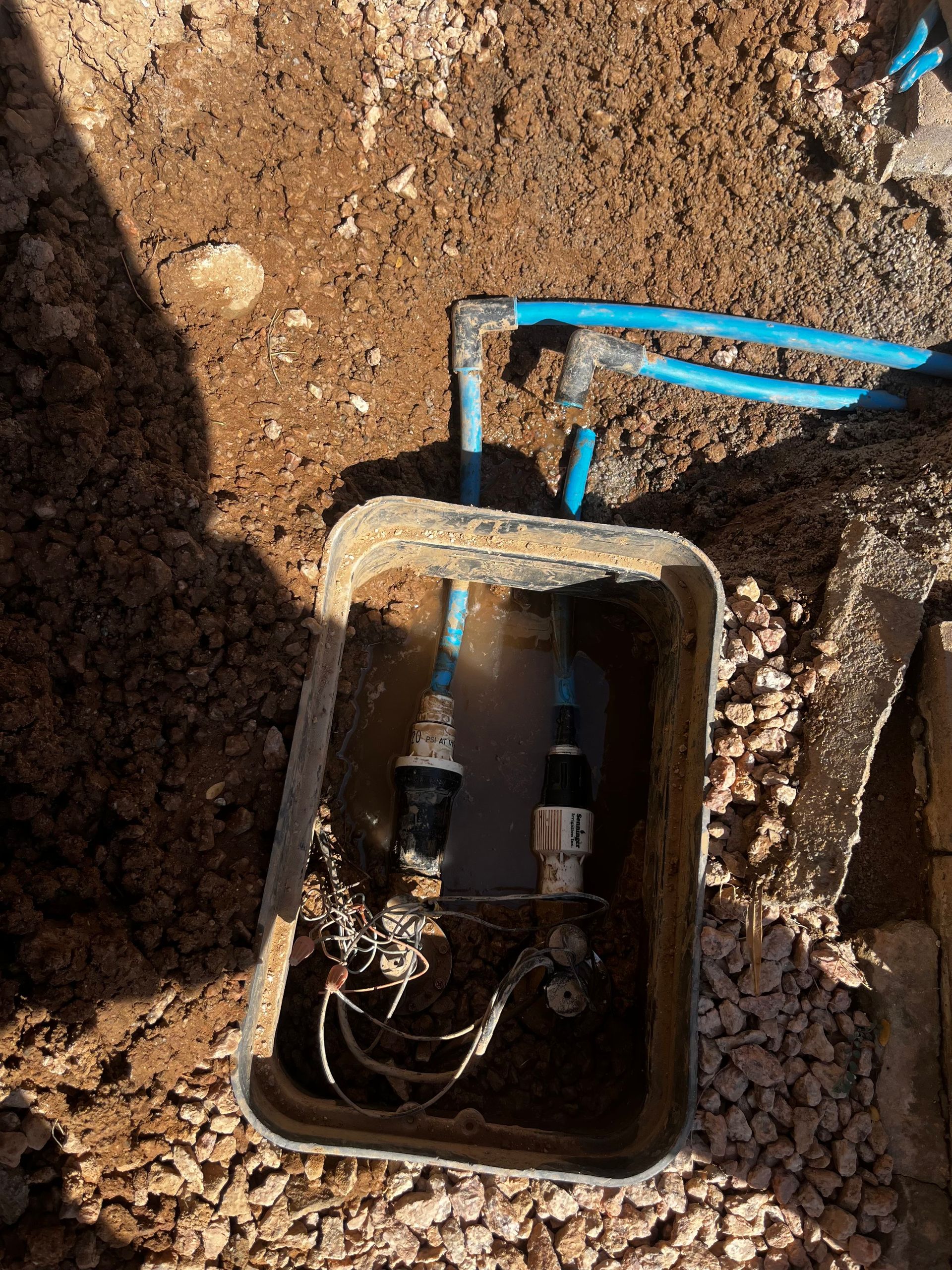 Open utility box in the dirt with blue pipes and black and silver connectors.