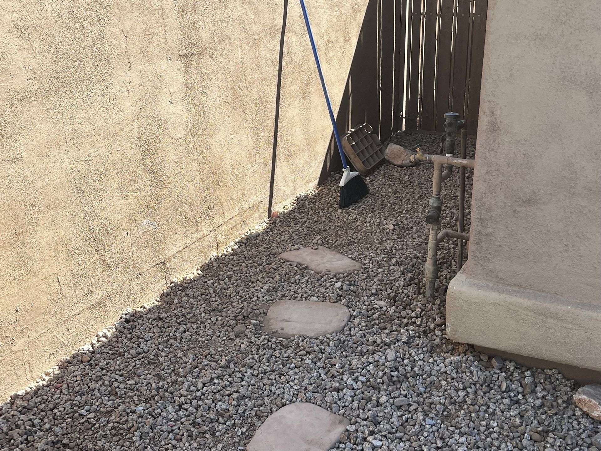 A stone pathway in a gravel bed alongside stucco walls and a wooden fence; broom leans against the wall.