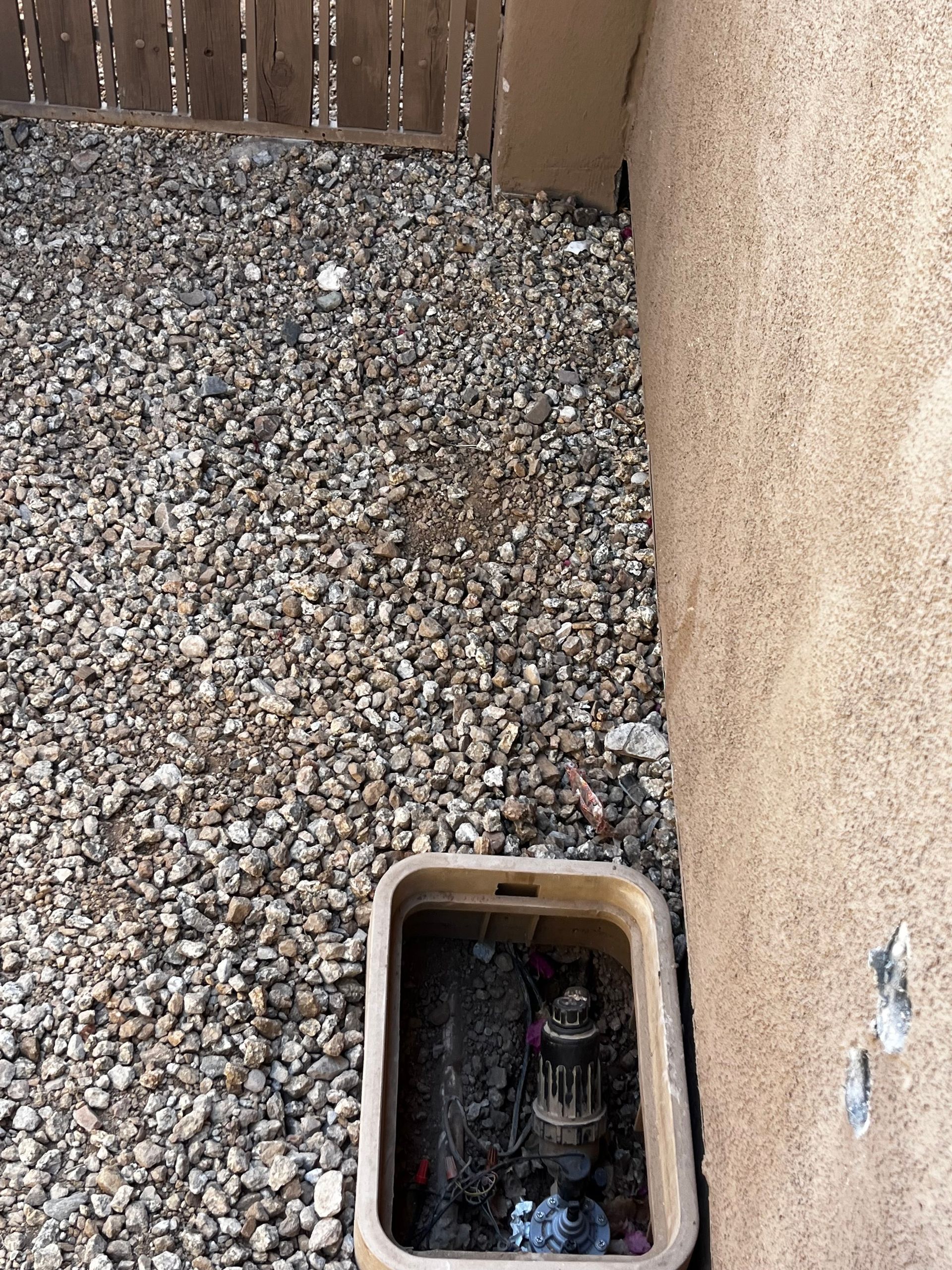 Brown water valve box in gravel next to a stucco wall and wooden fence.