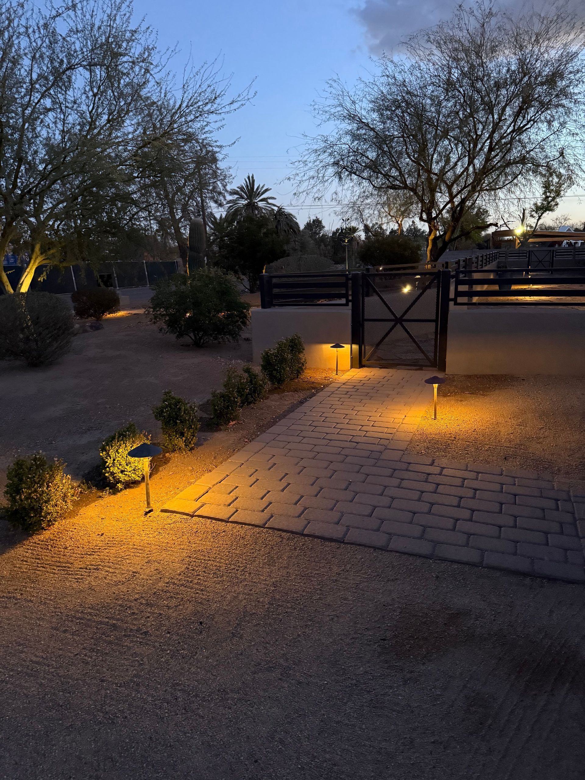 Pathway lit by warm lights, leading to a gate at dusk. Trees and shrubs in the background.