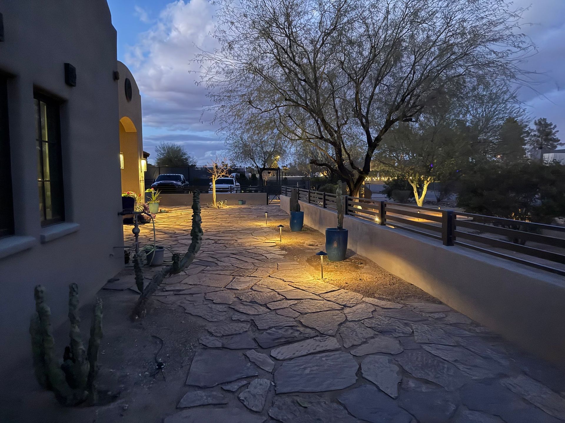 A stone path lit by spotlights at dusk leads through a desert landscape, with a house on the left.