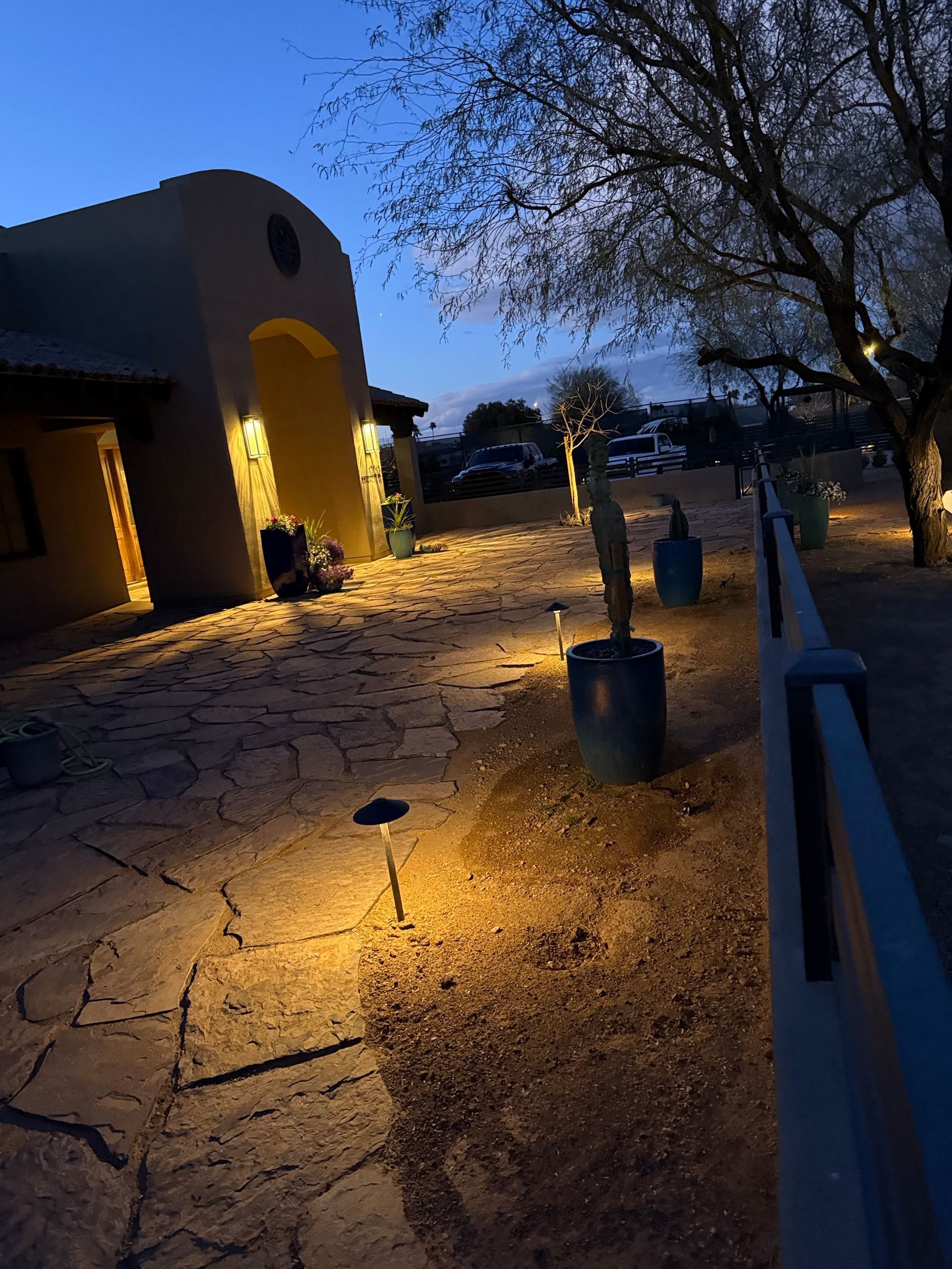 Path lit by spotlights leads to a building at dusk; cacti and trees line the path, blue sky.