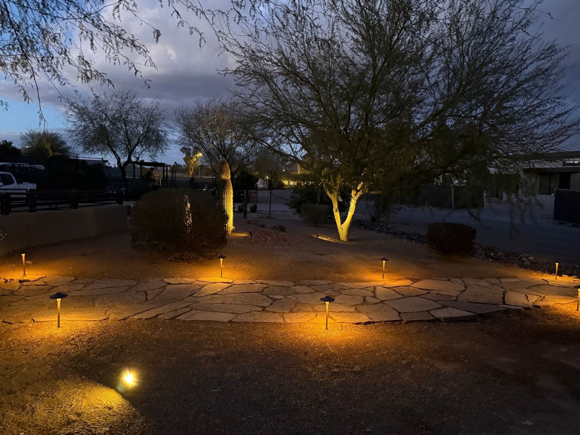 Outdoor path lit with warm lights at dusk, trees in the background.