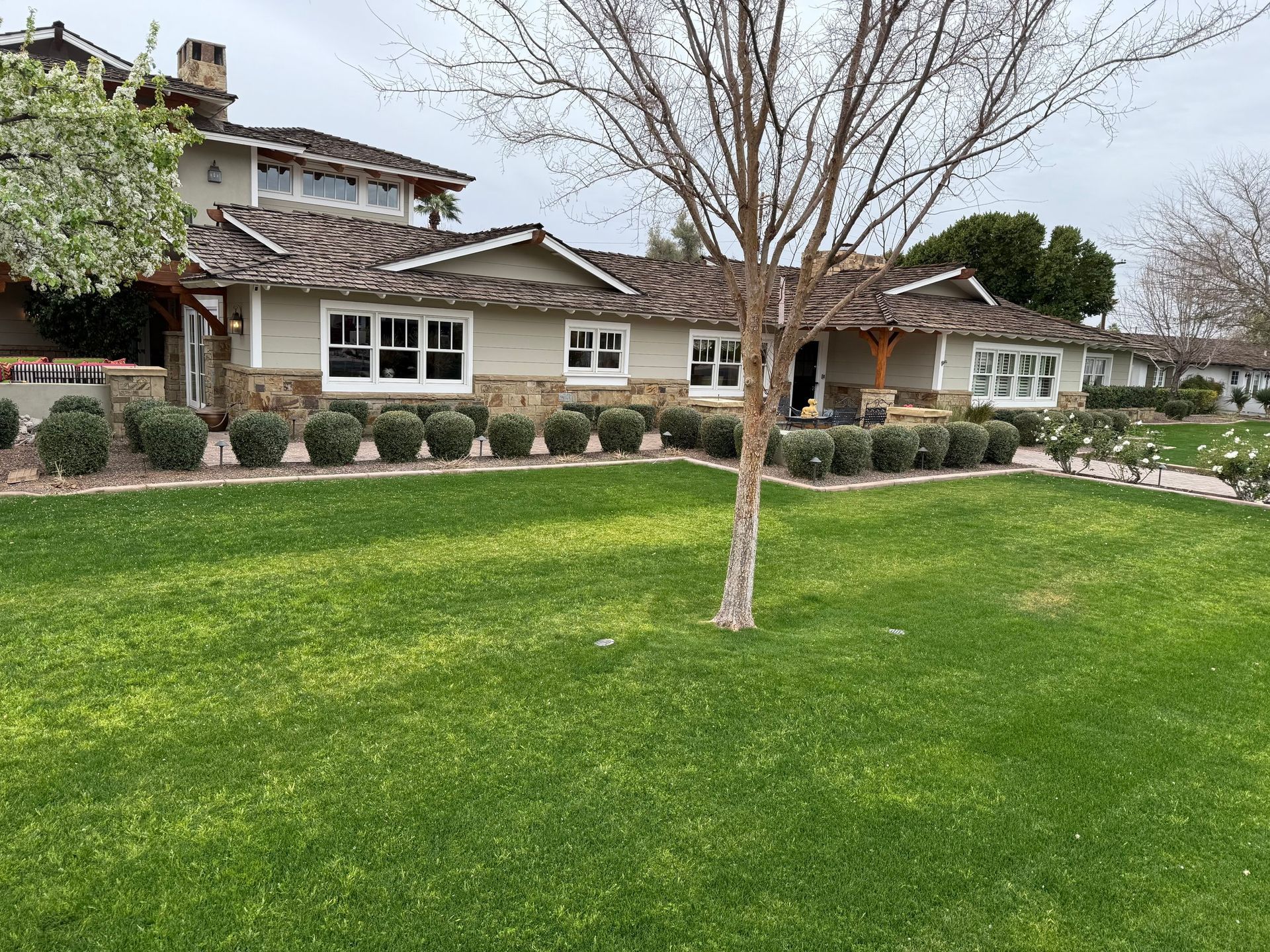 A single-story house with a light exterior, stone accents, and green lawn. Bushes line the front.