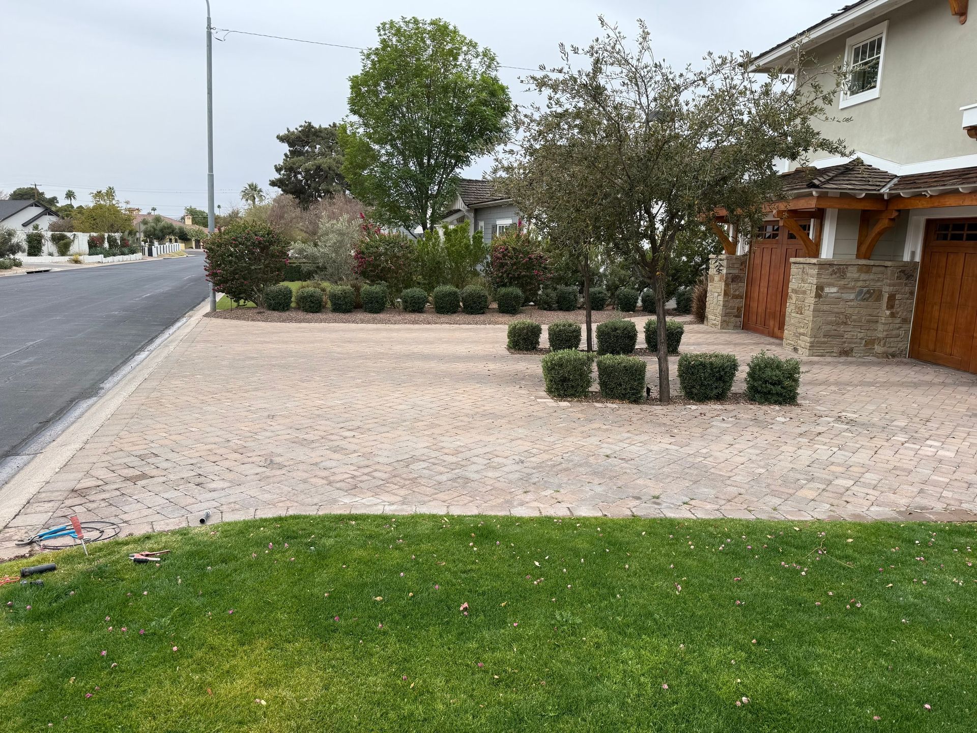 Driveway paved with stones in front of a house, bordered by bushes and lawn. Road and trees in background.