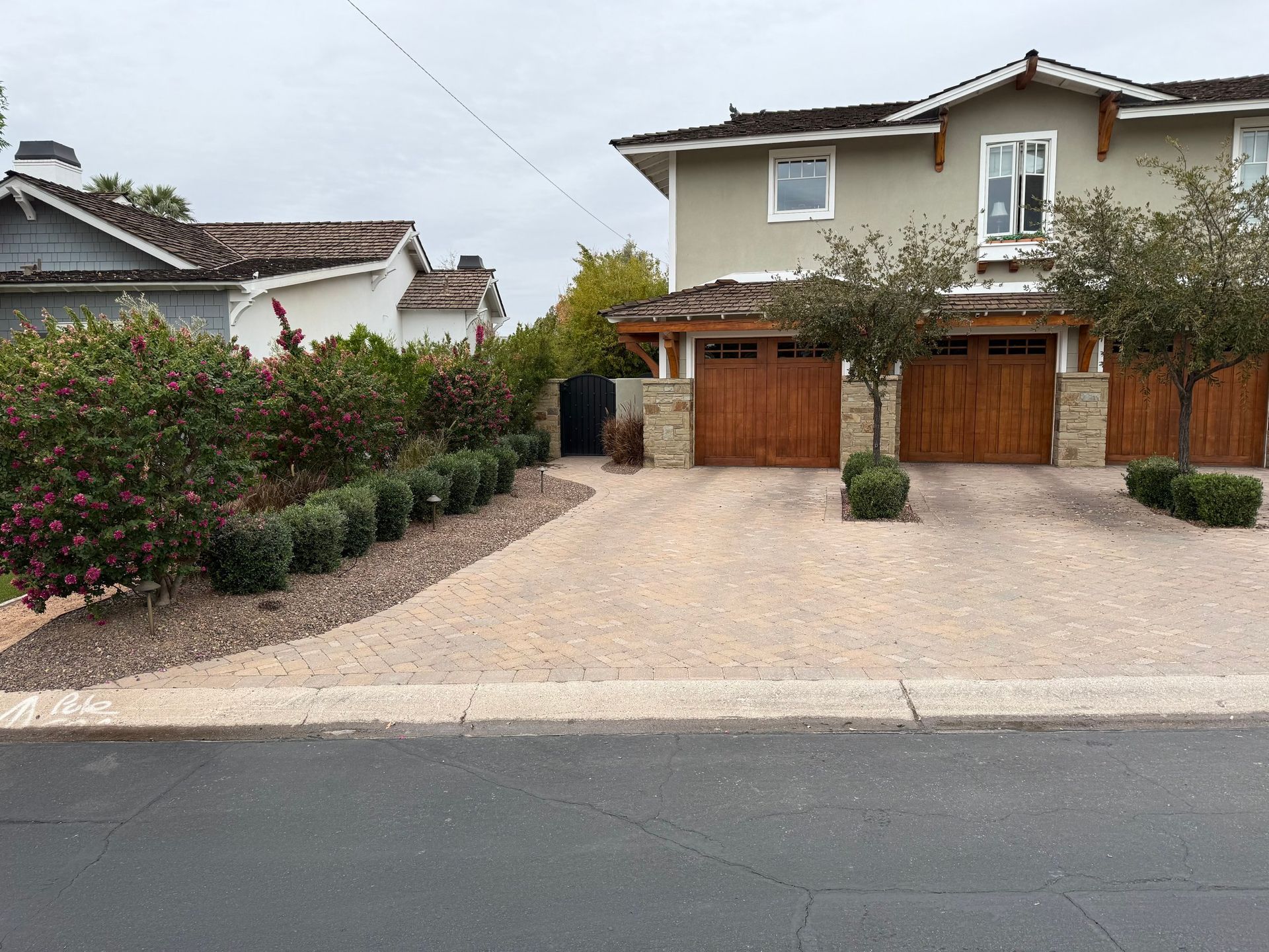 Two-story house with tan exterior and wooden garage doors. Driveway lined with shrubs and landscaping.