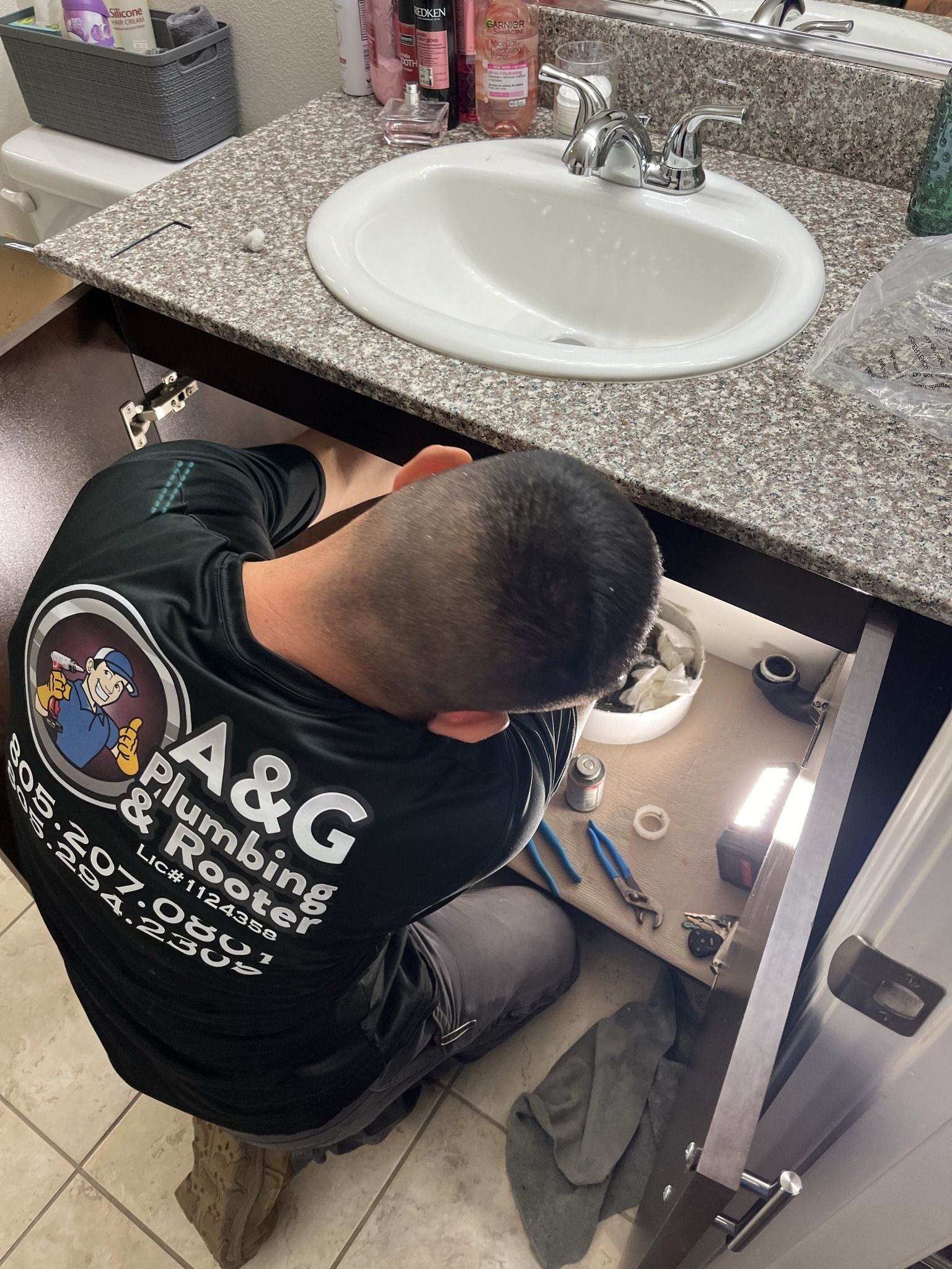 Plumber under a bathroom sink, working on pipes. The plumber wears a black shirt with company logo.