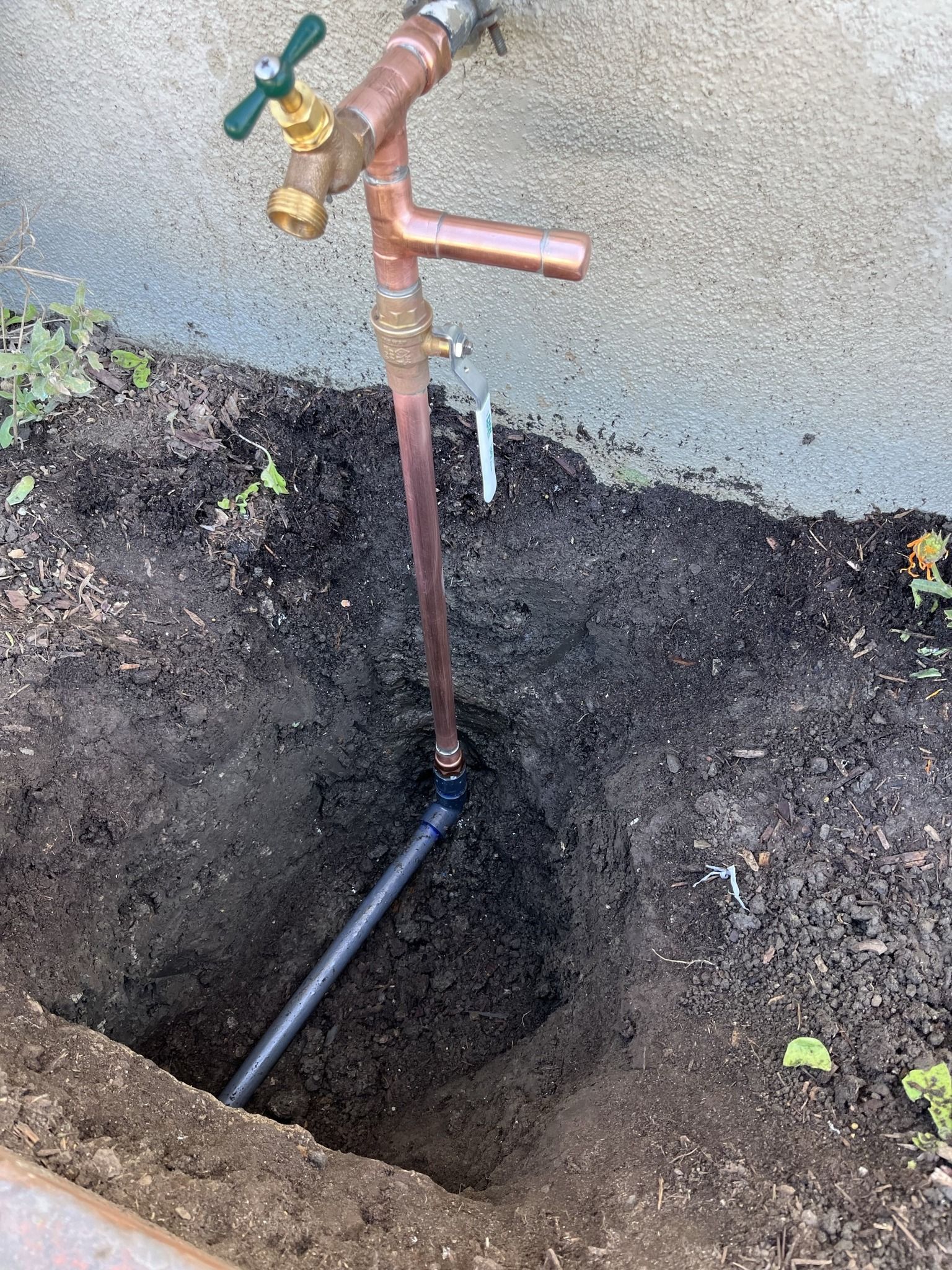 Copper water pipe exposed in a hole, leading to a spigot attached to a wall; soil surrounds.