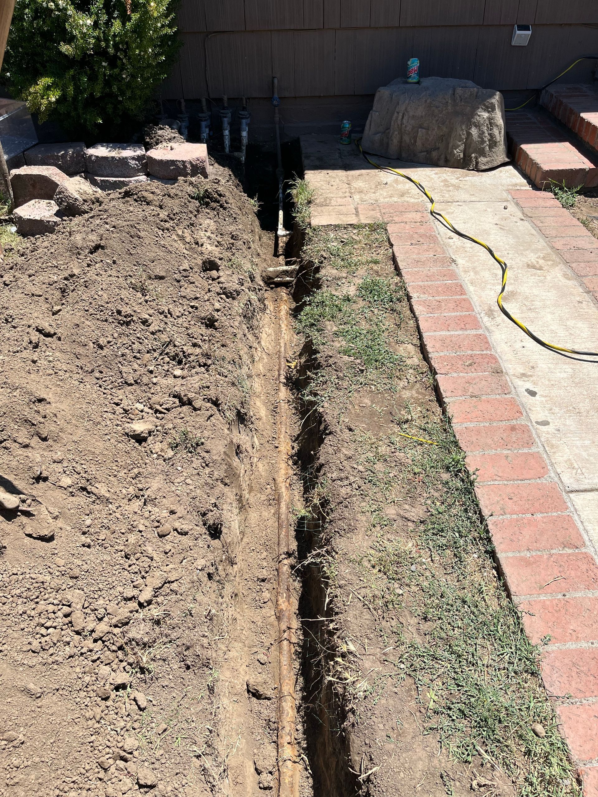 Narrow trench dug in brown soil alongside a brick pathway and grass.