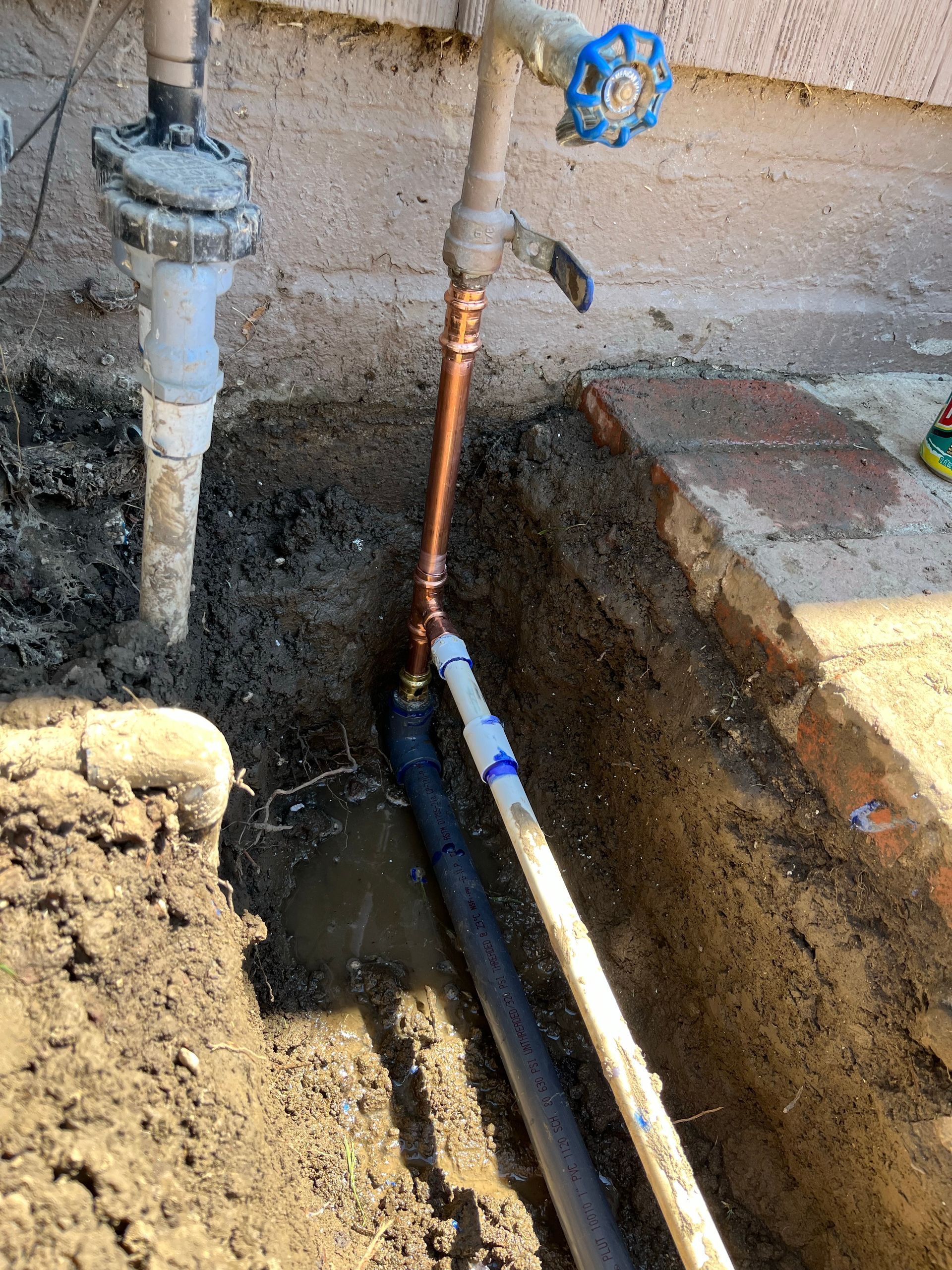 Trench with exposed copper and plastic pipes, a faucet, and water pooling, likely for irrigation repair.
