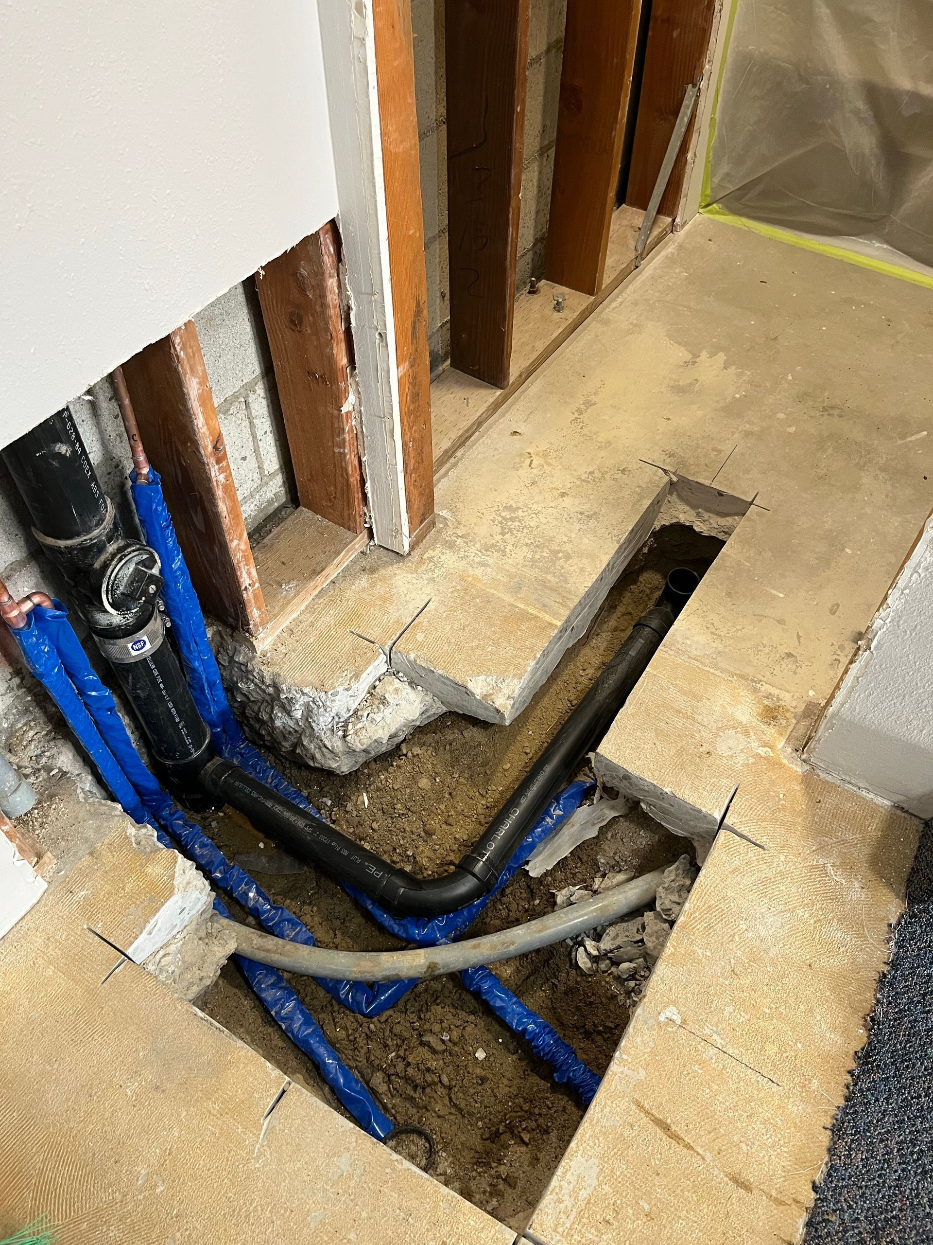 Plumbing work: black drain pipes and blue PEX tubing exposed in a floor opening surrounded by wood framing.