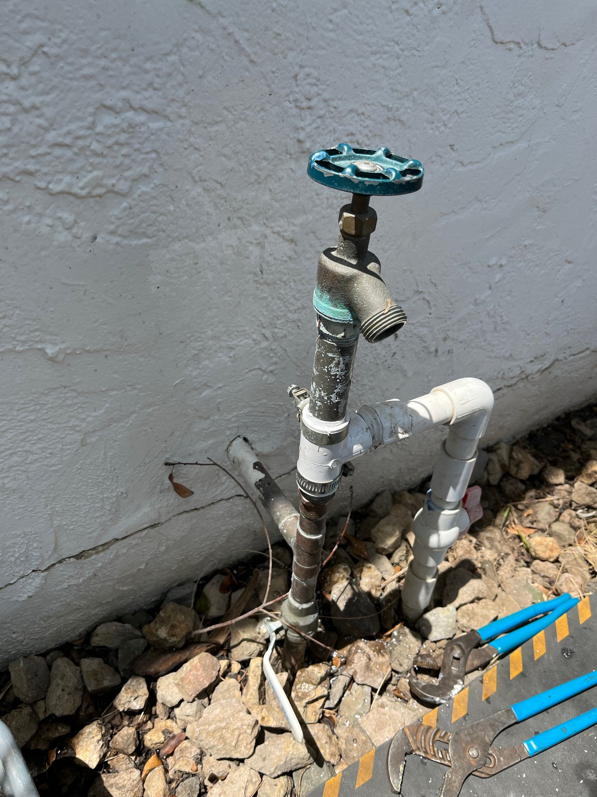 Outdoor water spigot against a white wall, surrounded by gravel. Spigot is teal, brown, and white.
