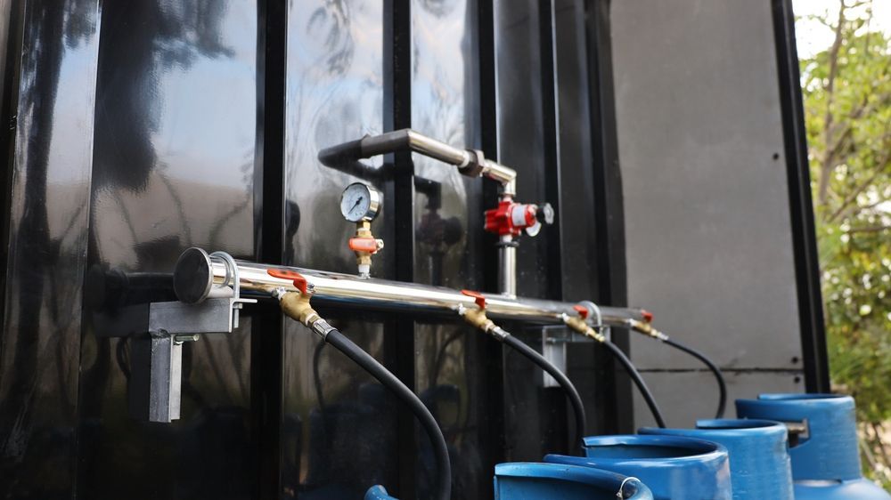 Black tank with pipes, gauges, and blue barrels below, likely for liquid storage.