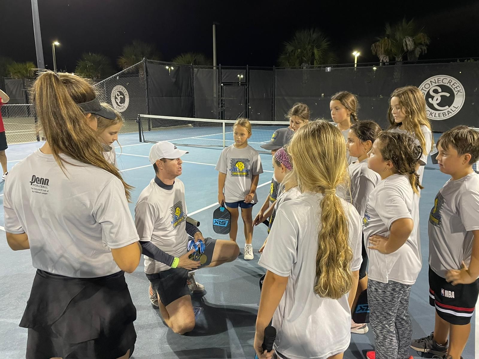 Group of children and adults on a pickleball court. Coach kneels, instructing the group wearing matching shirts.