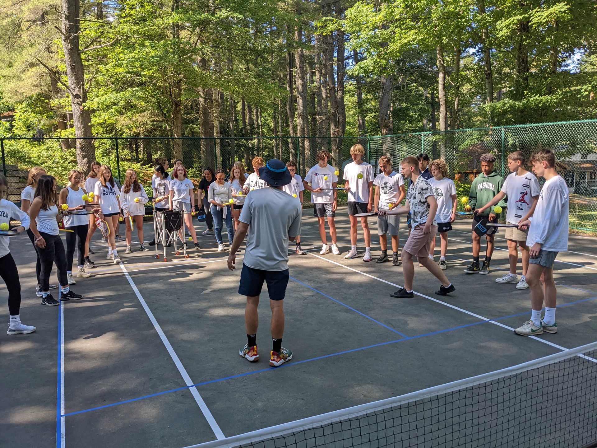A group of people on a pickleball court with a coach. Forest background.