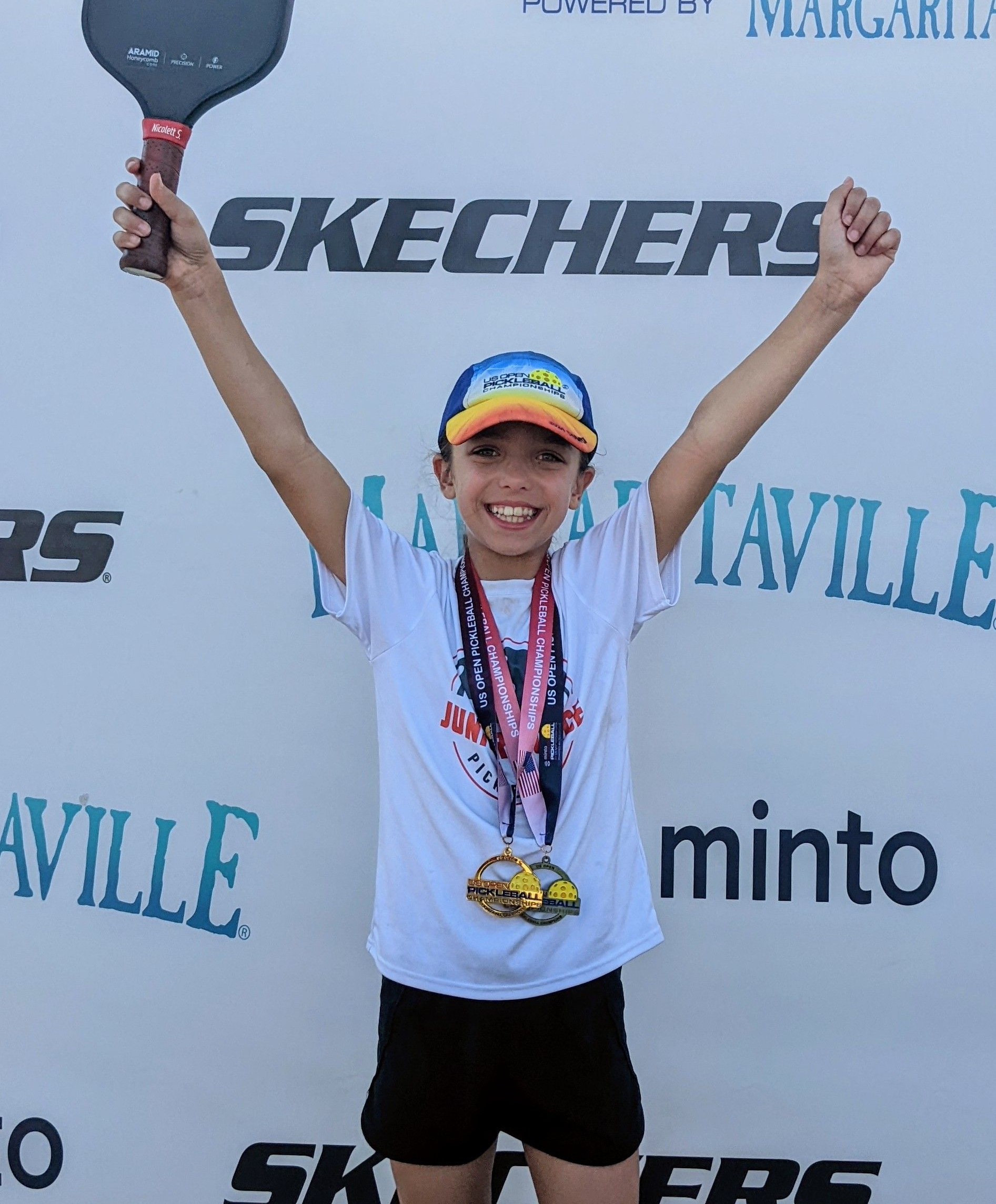 Person raises arms, holding paddle and wearing medal, celebrating a pickleball win in front of a sponsor backdrop.