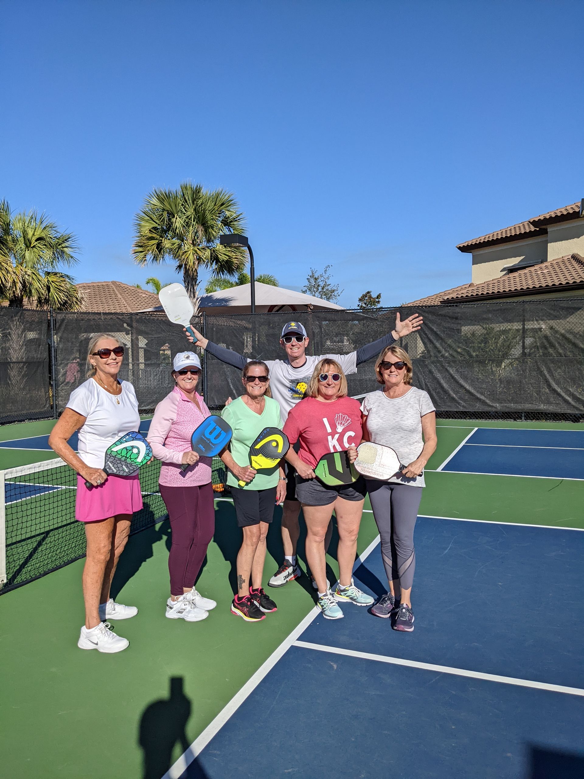 Group of six people on a pickleball court, holding paddles, posing for a photo, and smiling.