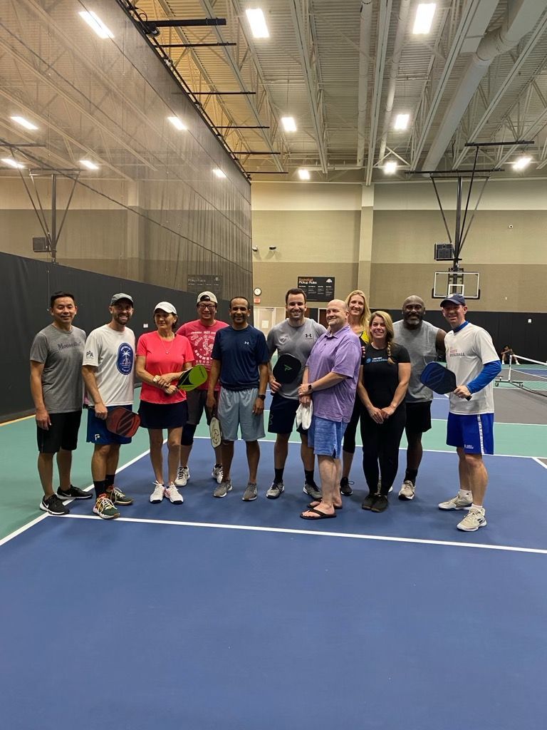 Group of people posing on a pickleball court, holding paddles. They are in an indoor sports facility.