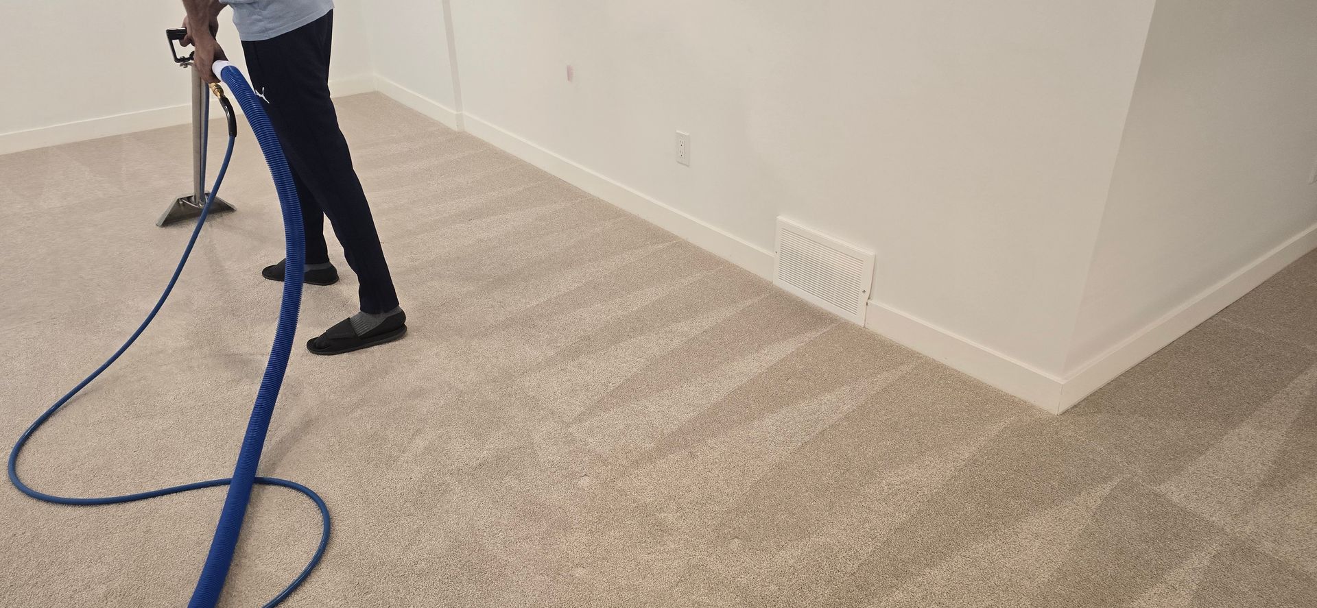 Person cleaning a beige carpet with a blue hose and a cleaning device in a room with white walls.