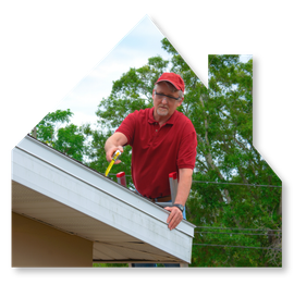 A person in a red shirt and cap measures the edge of a roof with a yellow tape measure outdoors against a background of trees.