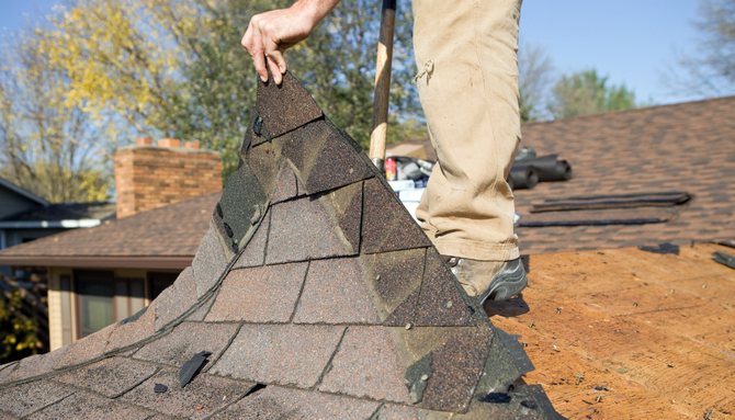 A person removing old asphalt shingles from a roof on a sunny day.