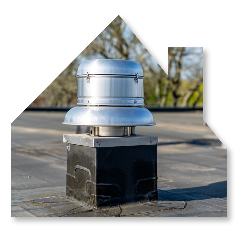 A metal roof exhaust vent mounted on a square black curb on a flat rooftop against a background of trees.