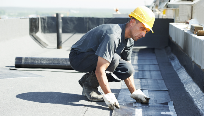 A worker in a yellow hard hat and gloves installs waterproof roofing material on a flat rooftop.